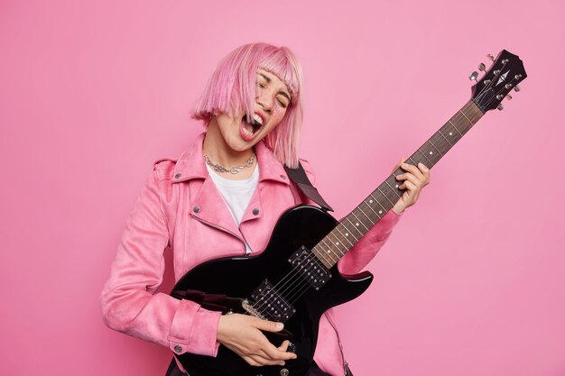 Сумасшедшая женщина играет хард-рок на электрогитаре, громко поет песню, демонстрирует свой талант, готовится к музыкальному фестивалю, принимает модные позы с розовой прической в помещении. Профессиональный гитарист или певец