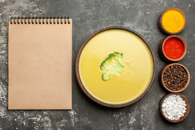 Creamy broccoli soup in a brown bowl and different spices next to notebook on gray table