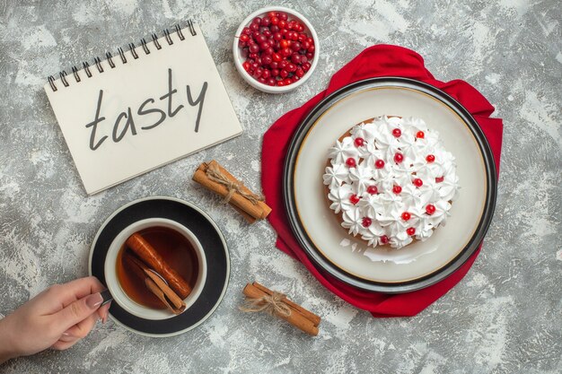 Сливочный торт, украшенный фруктами на красном полотенце и чашка черного чая с корицей, лаймом, смородиной, в белом горшочке рядом со спиральной записной книжкой на ледяном фоне