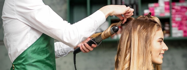 Создание локонов с помощью плойки. парикмахер делает прическу молодой женщине с длинными рыжими волосами в салоне красоты. Premium Фотографии