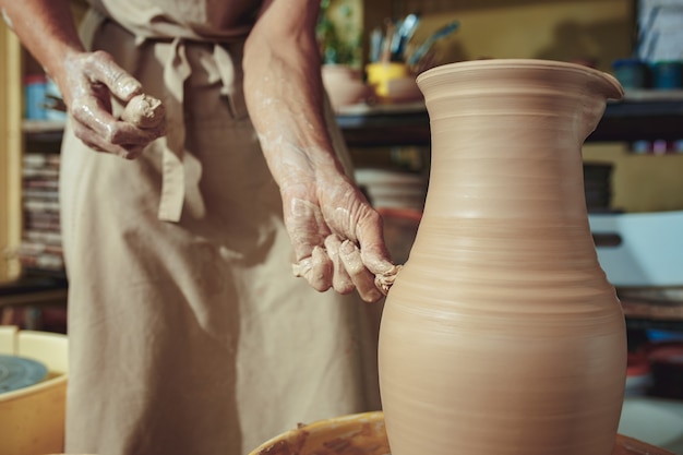 Free photo creating a jar or vase of white clay close-up. master crock. man hands making clay jug macro. the sculptor in the workshop makes a jug out of earthenware closeup. twisted potter's wheel.