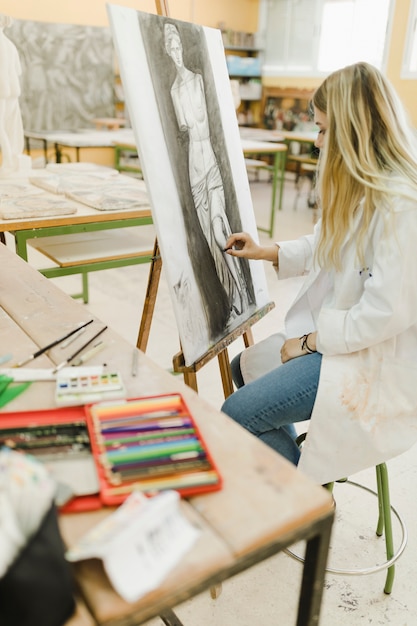 Creative artist sitting on stool sketching on easel with black crayon