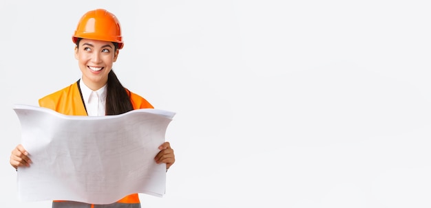 Architetto femminile asiatico creativo che immagina il suo piano in piedi nel casco di sicurezza e distoglie lo sguardo pensieroso mentre legge i progetti immaginando l'edificio dopo il completamento dei lavori di costruzione sullo sfondo bianco