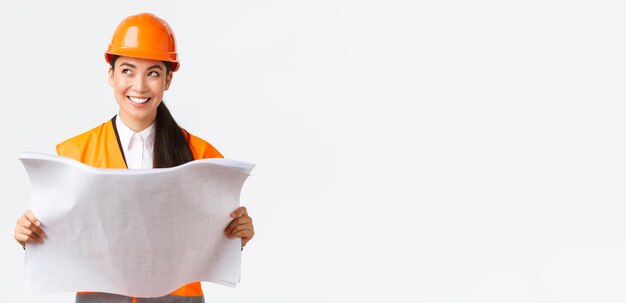 Creative asian female architect imaging her plan standing in safety helmet and looking away thoughtful while reading blueprints picturing buiding after finish construction works white background