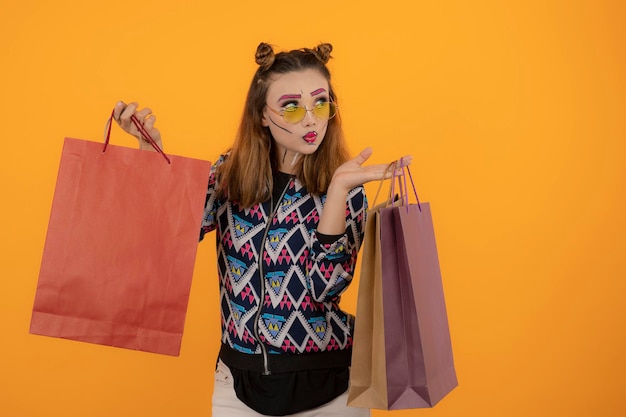 Креативная красивая девушка держит сумки с покупками и смотрит в сторону на оранжевом фоне. Фото высокого качества