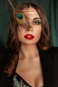 Творческий красочный портрет довольно молодой сексуальной женщины с ярким профессиональным макияжем и красными губами в модной черной одежде закрывает глаза павлиньим пером на винтажном зеленом фоне