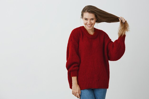 Creative coworker flirting with handsome guy during break. Studio shot of good-looking feminine european woman in loose red sweater, pulling hair and smiling broadly, taking good care about health