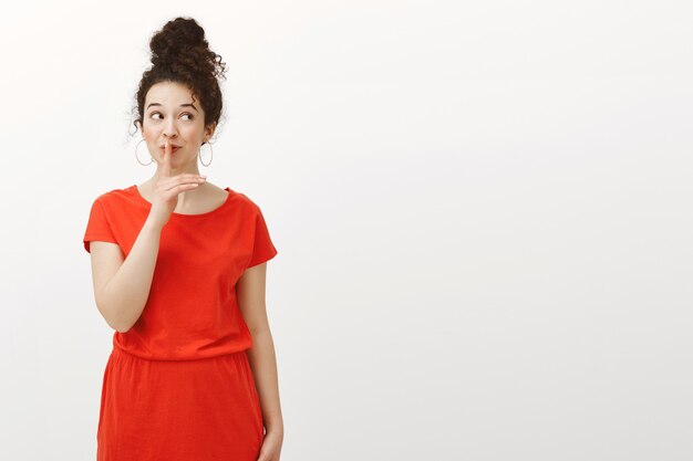 Creative emotive happy woman with curly hair in stylish red dress, looking right and smiling broadly while saying shh