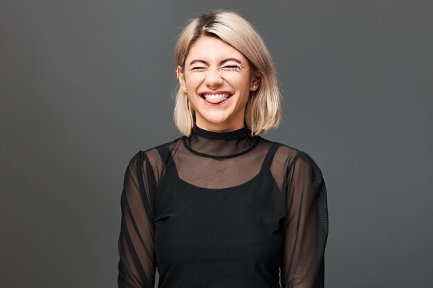 Creative fashion mascarade and Halloween celebration. Elegant flirty cheerful young woman with short blonde hair and artistic make up laughing out loud posing at dark wall in fancy black dress