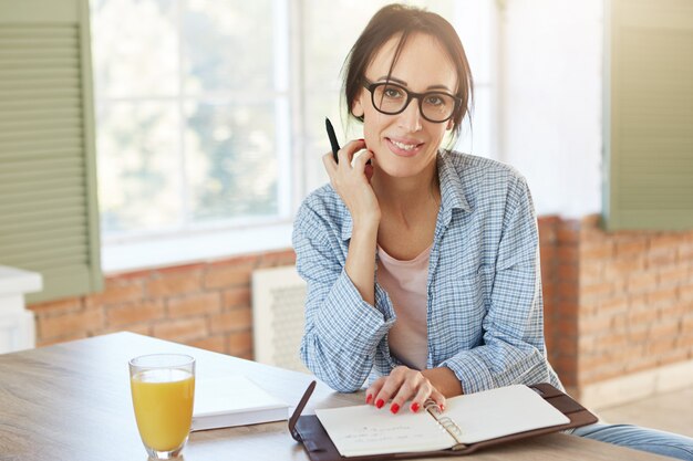 Creative female worker being at home, writes notes and plans her schedule, looks into camera. Woman freelancer works remotely, sits in kitchen.