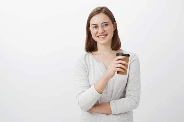 Творческая счастливая и энергичная привлекательная молодая женщина в очках держит бумажный стаканчик, пьет кофе и посмеивается, весело и забавно разговаривая во время обеда на работе над серой стеной