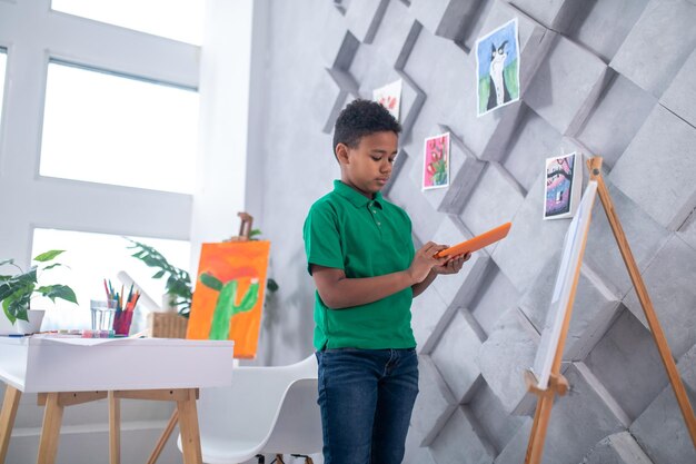 Creative mode. Concentrated dark-skinned school-age boy in green t-shirt and jeans looking at tablet near drawing easel in light room with drawings on wall