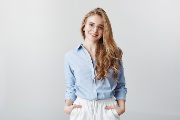 Creative office worker relaxing after long day at work. Positive good-looking female with blond hair in blue blouse, holding hands in pockets and smiling, expressing confidence over gray wall