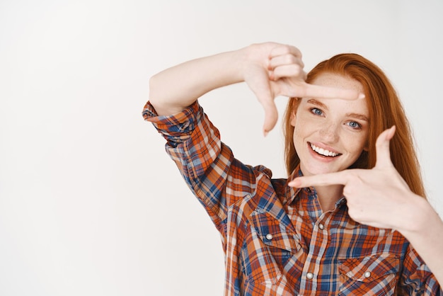 Free photo creative redhead girl making camera hand frames and looking happy capturing moment standing over white background