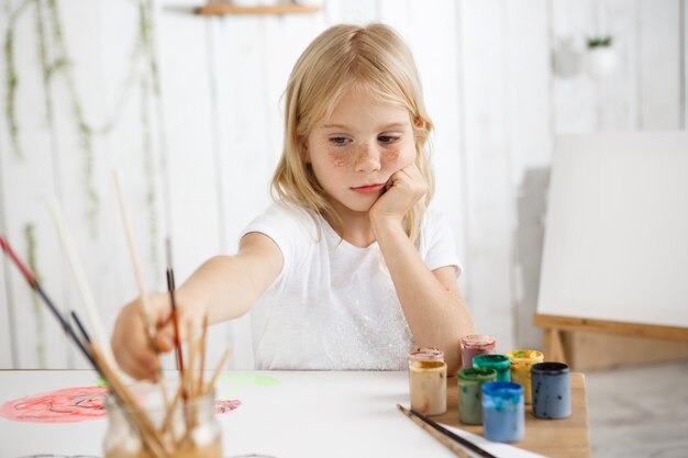 Творческая семилетняя девочка рисует акварелью, сидит за столом и кладет локти на стол