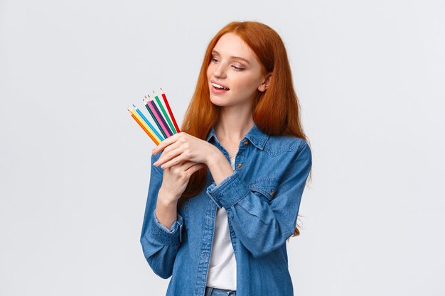 Креативная и умелая красивая рыжая женщина в джинсовой рубашке, выбирает цветные карандаши, улыбается, думая, что рисовать, создает произведения искусства, задумчиво стоит на белом фоне.