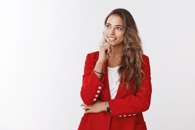 Creative skillful female employee thinking ideas working important project looking up thoughtful satisfied smiling touching jawline thinking, having plan, standing dreamy wearing red formal jacket
