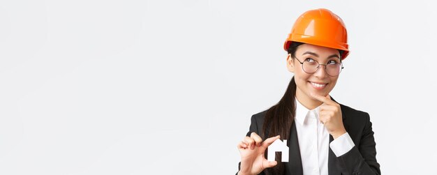 Креативная улыбающаяся азиатская женщина-архитектор думает о новом дизайне во время строительных работ, выглядя задумчивой с улыбкой, держа в руках миниатюрный домик на белом фоне