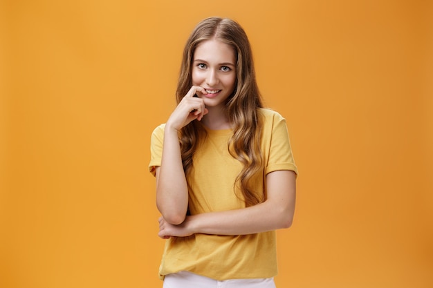 Creative tricky young woman with natural wavy long hair in yellow t-shirt looking from under forehead with intention and lust in expression biting finger, smiling at camera over orange wall.