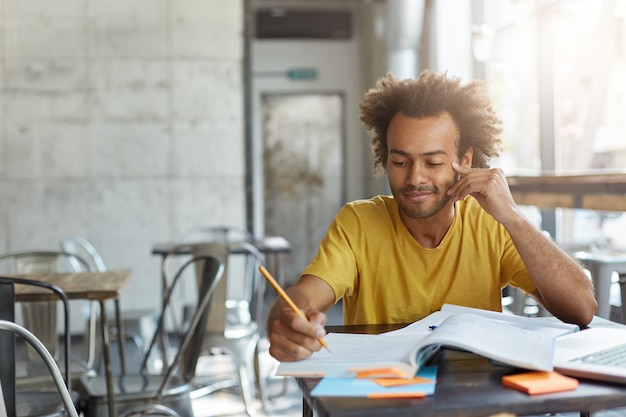 Free photo creative young author with curly hair and dark skin dressed casually sitting at cafeteria preparing for writing new article in his newspaper having gentle smile on his face having good ideas in mind