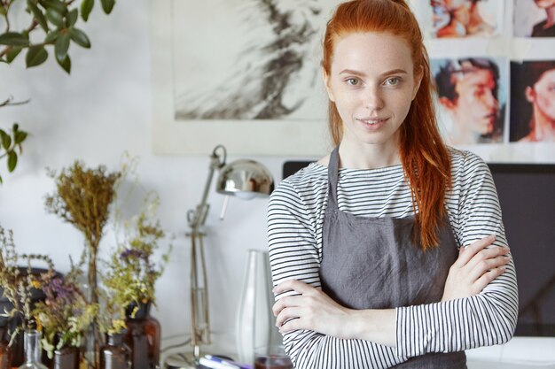 Creativity and art concept. Waist up shot of beautiful creative professional woman artist with long ginger hair standing in workshop space with pictures on wall and flowers in bottles on table