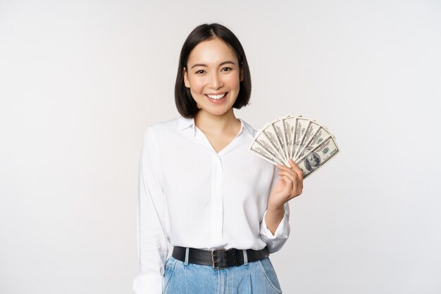 Concetto di credito e prestito sorridente giovane donna asiatica in possesso di dollari in contanti e guardando felice lo sfondo bianco della fotocamera