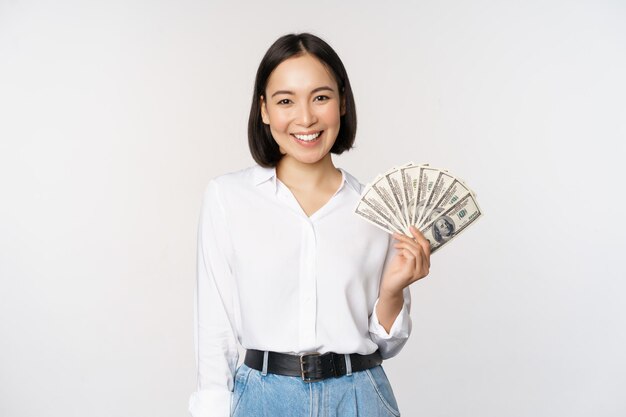 クレジットとローンの概念現金ドルを保持し、カメラの白い背景で幸せそうに見える若いアジアの女性の笑顔