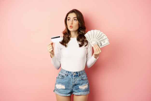 Credit, money and loans concept. Happy beautiful girl holding credit card and cash, looking satisfied, thinking of shopping, standing over pink background.
