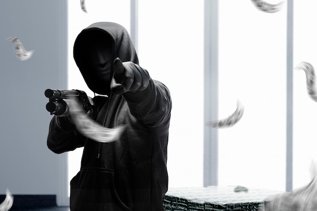Преступник в скрытой маске держит дробовик и что-то направляет во время грабежа денег на банке.