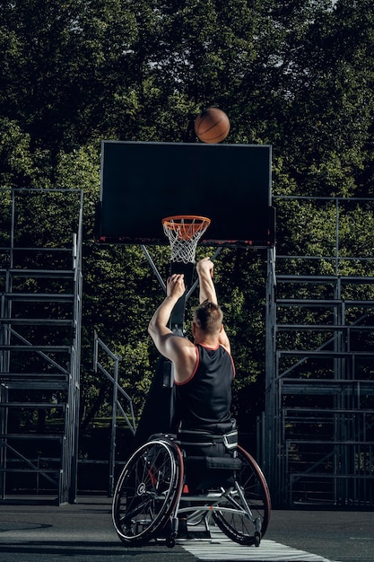 Cripple basketball player in wheelchair plays basketball on open air ground.