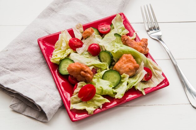 Салат из хрустящей курицы с попкорном, украшенный овощами в красной тарелке с салфеткой и вилкой