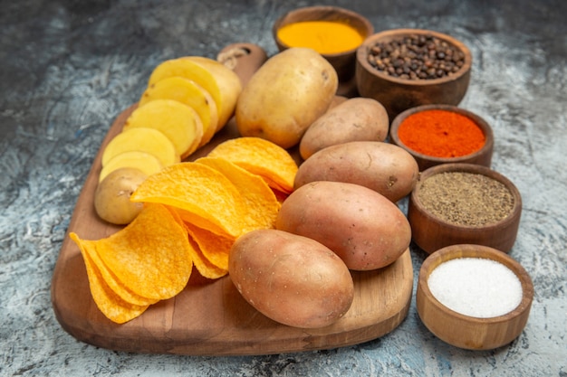 Crispy chips and uncooked potatoes on wooden cutting board and different spices on gray table