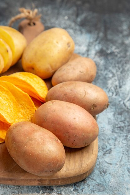 Crispy chips and uncooked potatoes on wooden cutting board on gray table