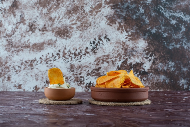 Бесплатное фото Хрустящие картофельные чипсы и йогурт в тарелках на подставках, на мраморном столе.