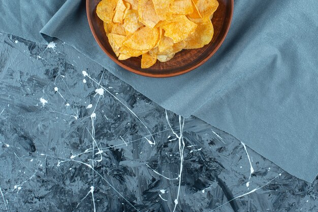 Crispy potato chips in plate on pieces of fabric on marble.