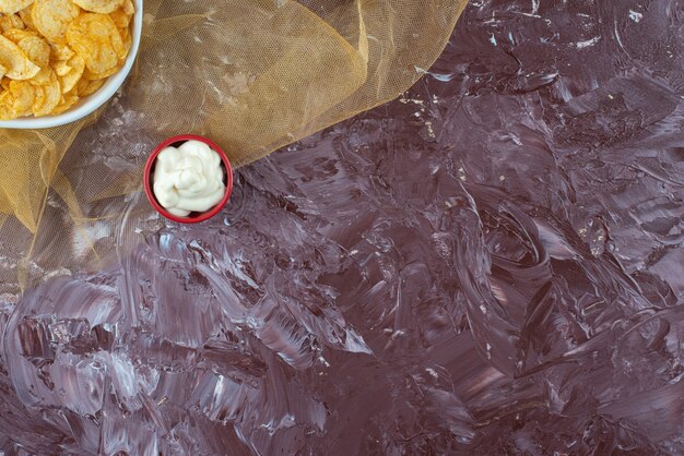 Хрустящие картофельные чипсы и йогурт в тарелках на тюле, на мраморном столе.