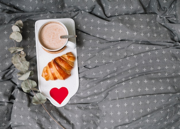 Croissant, cuore dell'ornamento e tazza di bevanda sul tagliere vicino al ramoscello