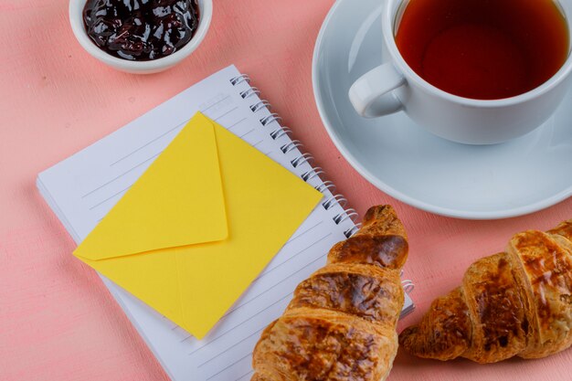 Croissant with tea, jam, envelope, notebook close-up on a pink table