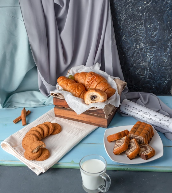 Croissants, sliced pie and cookies with a cup of milk on the blue wooden table.