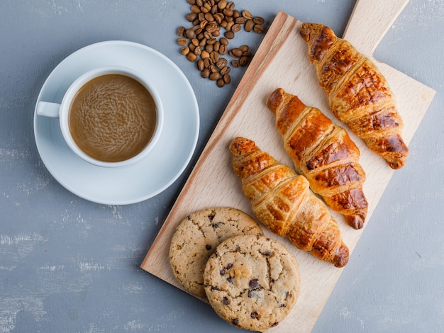 Croissants with coffee and beans, cookies on plaster and cutting board, flat lay.
