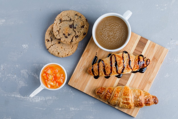 Croissants with coffee, cookies, sauce flat lay on plaster and wooden board