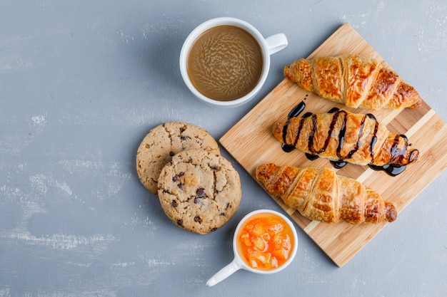 Croissants with coffee, cookies, sauce flat lay on plaster and wooden board