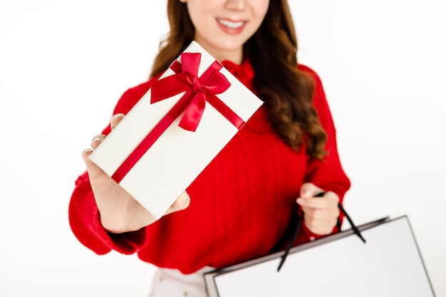 Урожай крупным планом азиатская женщина в красном платье с шляпой санты держит хозяйственные сумки и показывает подарочную коробку на белом изолированном фоне. красивая дама с длинными черными волосами. концепция покупок.