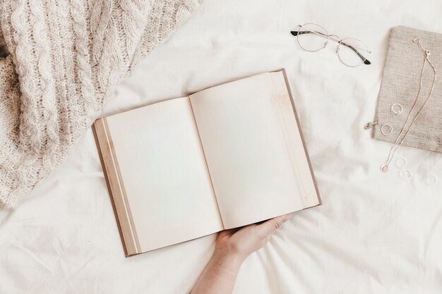 Crop hand holding book near accessories on bed