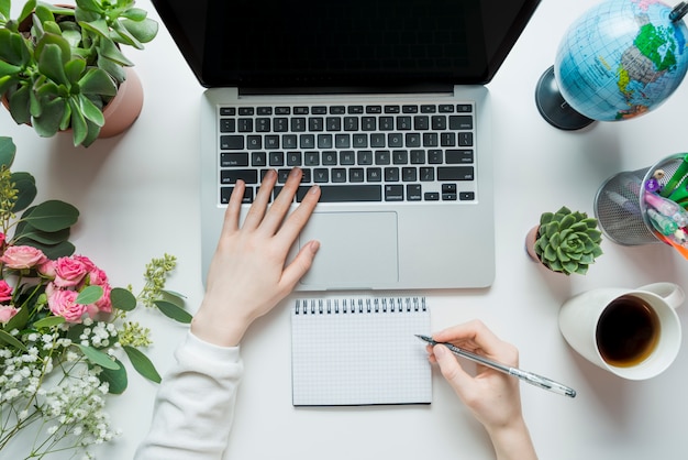 Crop hands making notes and using laptop