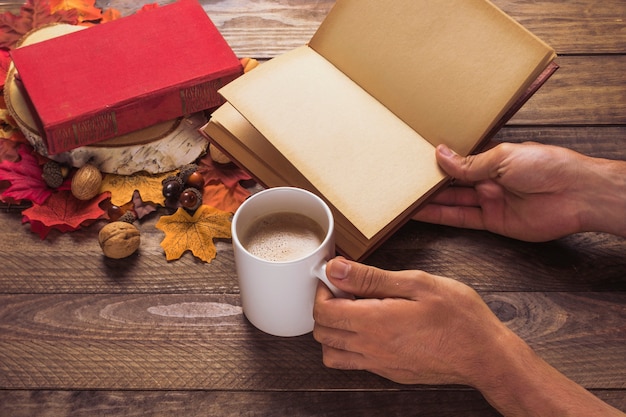 Crop hands with book and coffee near leaves and nuts