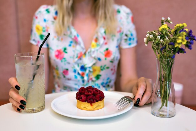 レストランでラズベリーのデザートを食べ、大きな甘いレモネードを持って、花柄のドレス、ピンクの背景、パステルカラーを身に着けている女性の作物画像