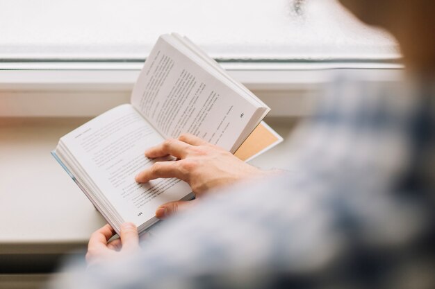 Crop man reading book near window