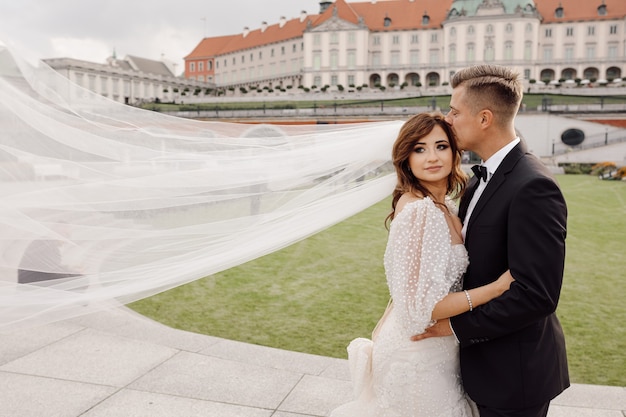 ウェディングドレスと白い長いベールでカメラを見て、笑顔で、結婚式の日は一緒に幸せです。背景に古いヨーロッパの都市 Premium写真