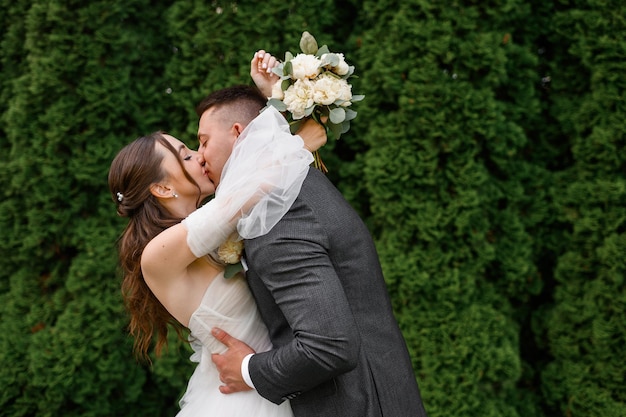 Ritaglia il ritratto delle coppie di spose amorevoli che si baciano e si abbracciano mentre si è in piedi in giardino bella ragazza brode con l'acconciatura riccia in abito da sposa con lo sposo in abito giorno del matrimonio momento di tenerezza